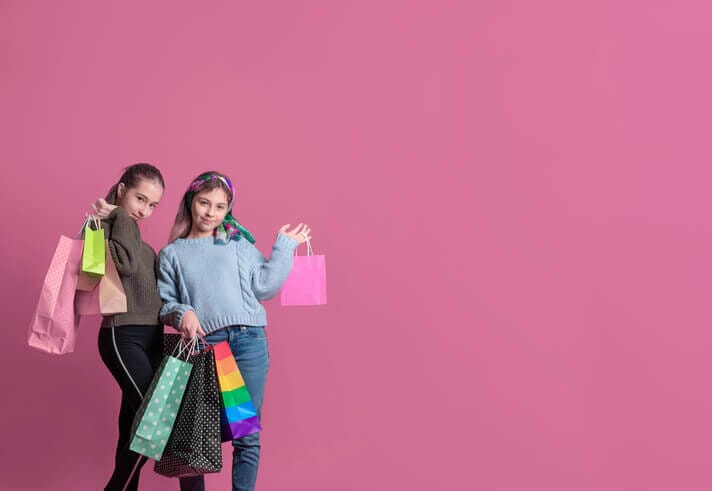 Two tweens holding shopping bags, posing with a playful attitude against a pink background. Image from iStock Photo by sonreir es gratis.