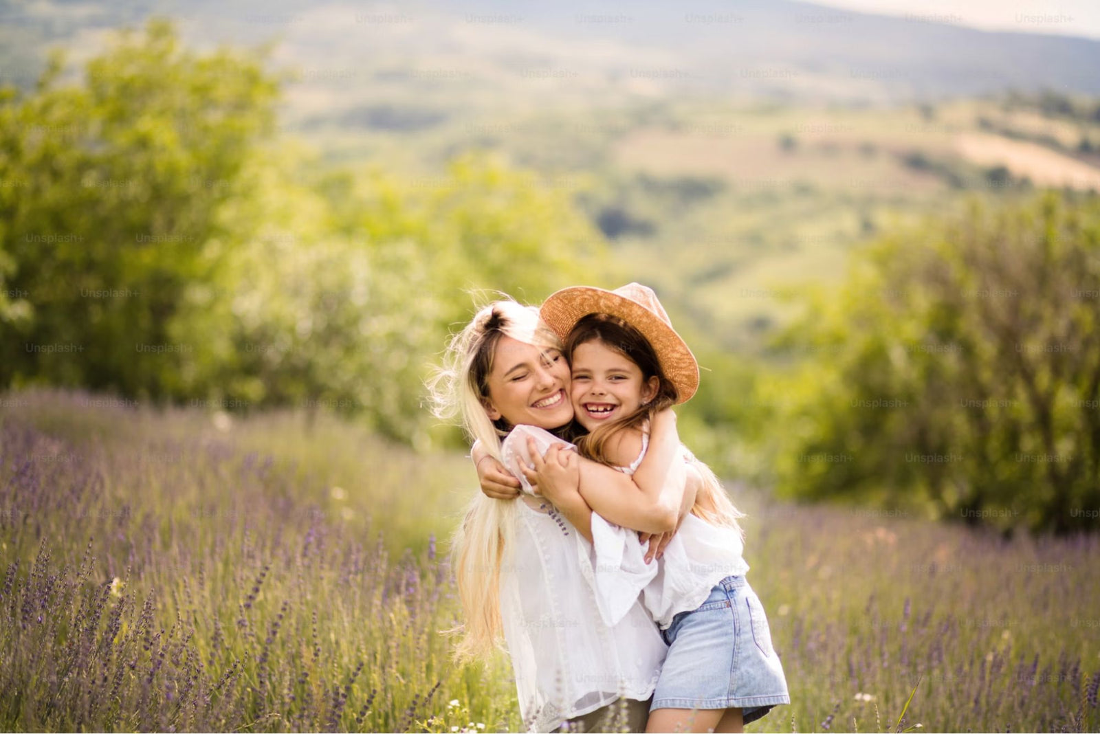 Smiling mother and young daughter hugging in a sunny lavender field, with the daughter wearing a straw hat and denim shorts. The background features green hills and a bright, cheerful atmosphere.