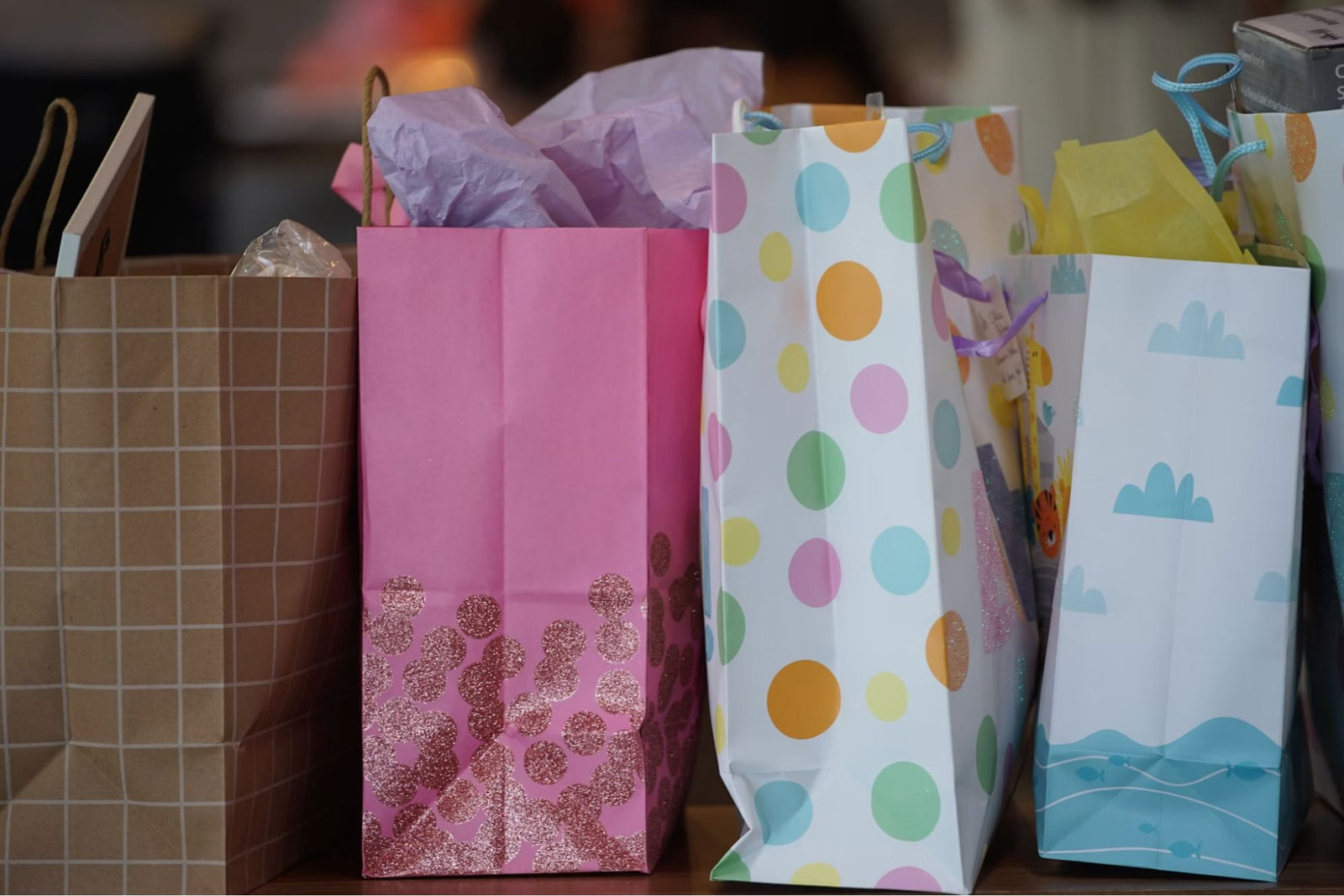 Row of colorful gift bags—brown grid, pink with glitter dots, white with pastel polka dots, and white with blue cloud-and-wave design—lined up on a table.
