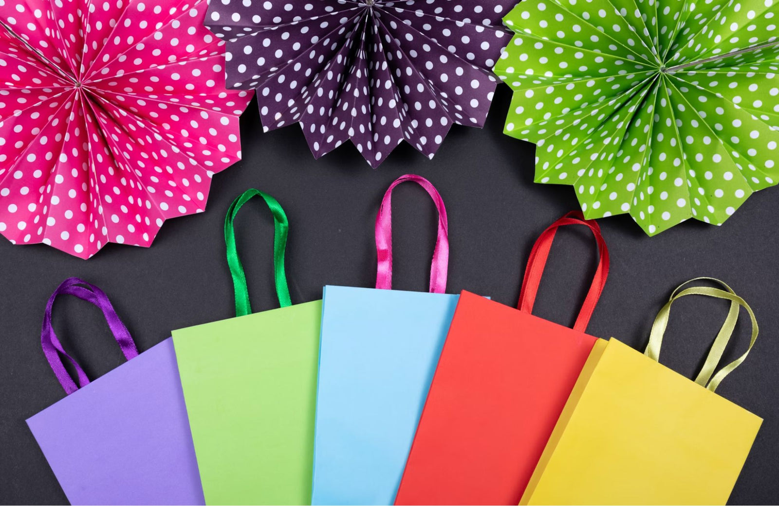 Colorful shopping bags with satin handles arranged in a row on a dark background, with vibrant polka-dotted paper fans in pink, purple, and green at the top for a festive, celebratory look.