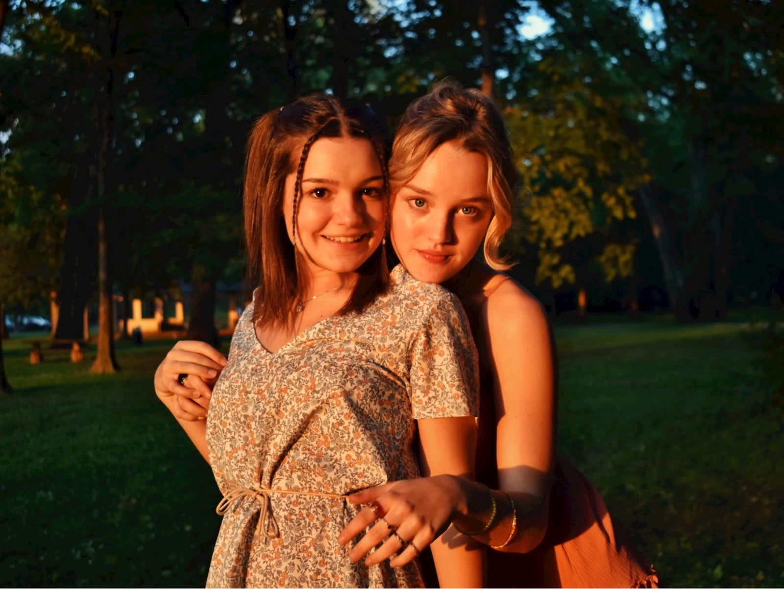 Two teen girls standing closely together in a sunlit park, wearing summer dresses and smiling at the camera
