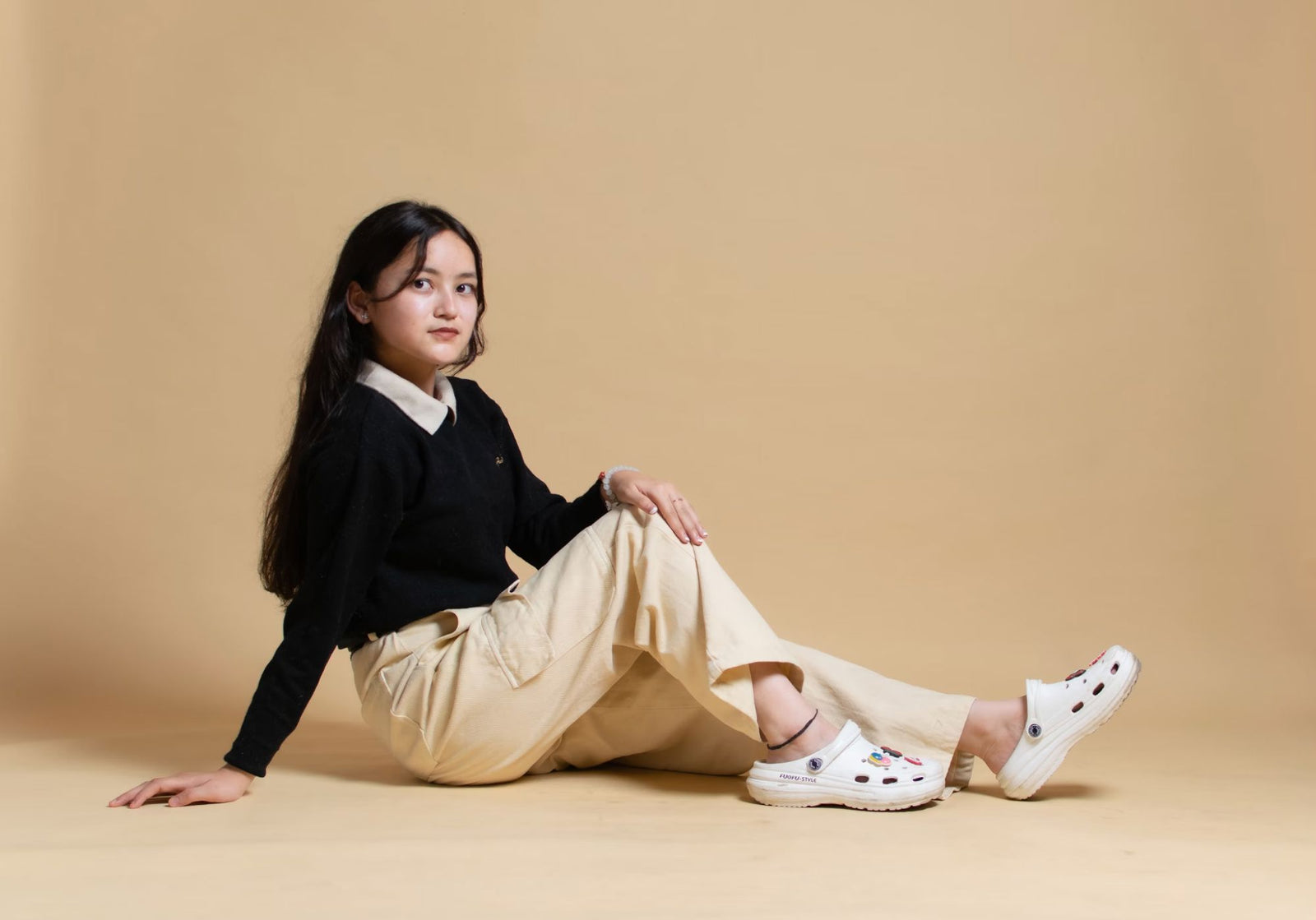 Teen girl seated on beige floor, in black sweater, cargo pants, and white Crocs.