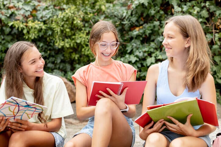 Three young teen girls, each holding a notebook, are sitting together and smiling, with the central girl in glasses reading from her red notebook. Image from iStock Photo by Alina Rosanova.