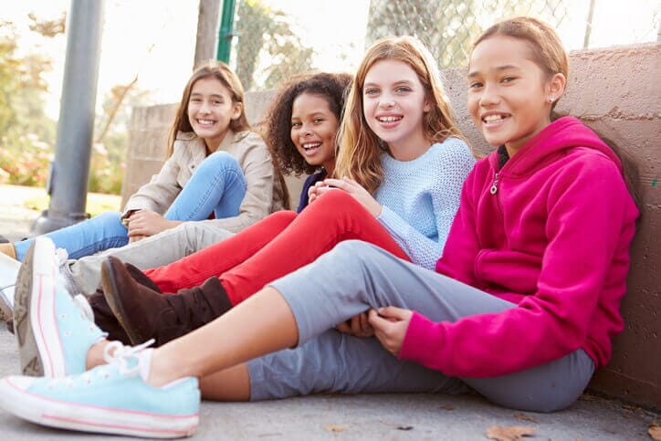 Four girls in casual attire sitting on the ground leaning against a wall, smiling at the camera. Image from iStock Photo by monkeybusinessimages.