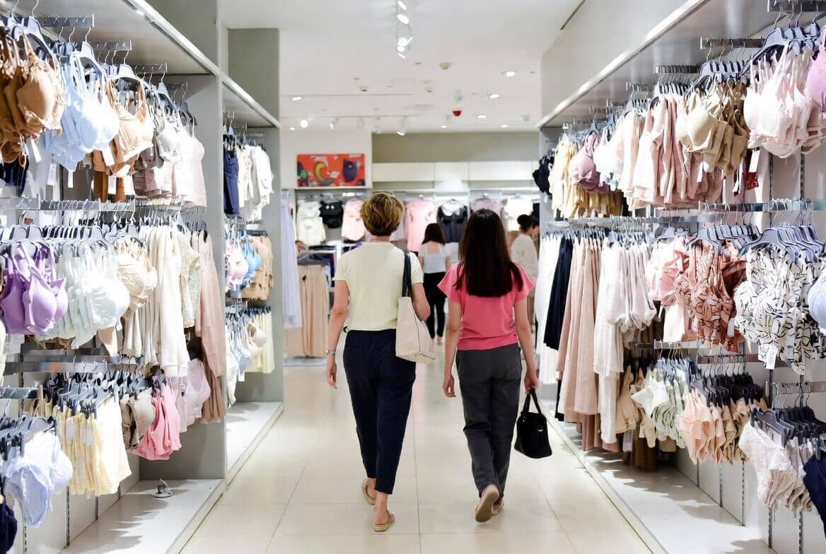 A mother and her teenage daughter shopping together in a lingerie store aisle, both holding shopping baskets and examining underwear items 