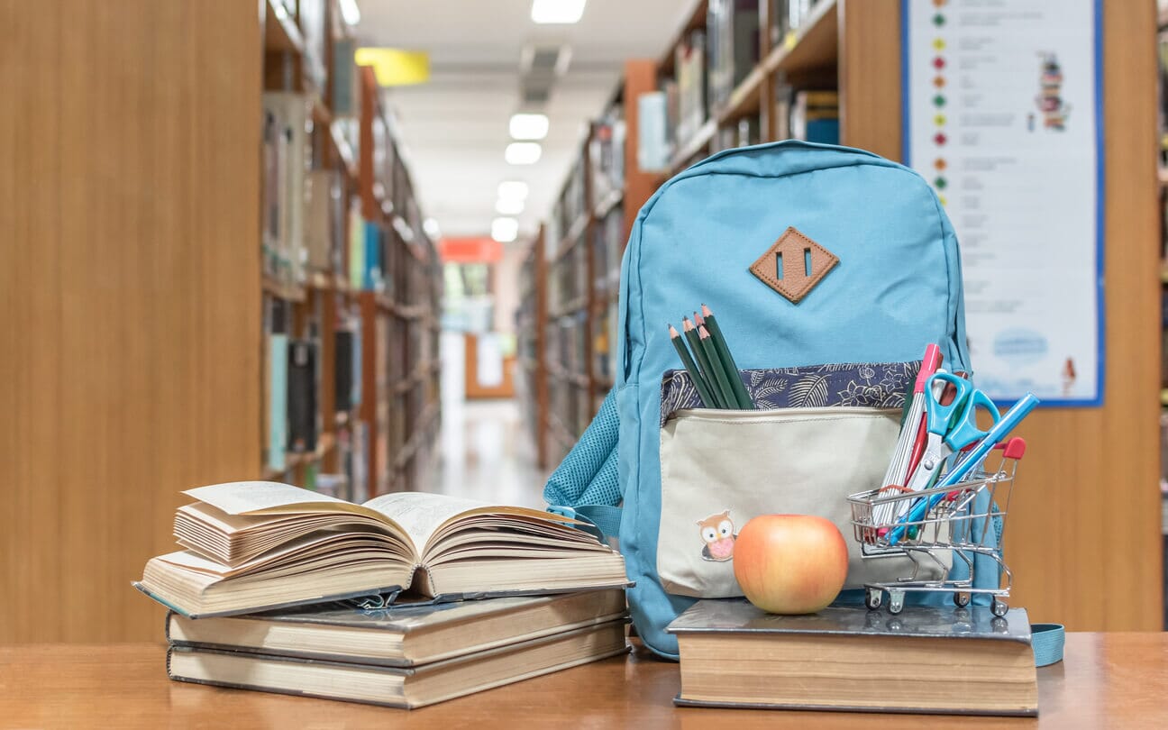 A back-to-school concept image with books, a blue backpack, school supplies and an apple on a classroom desk with a library in the background.