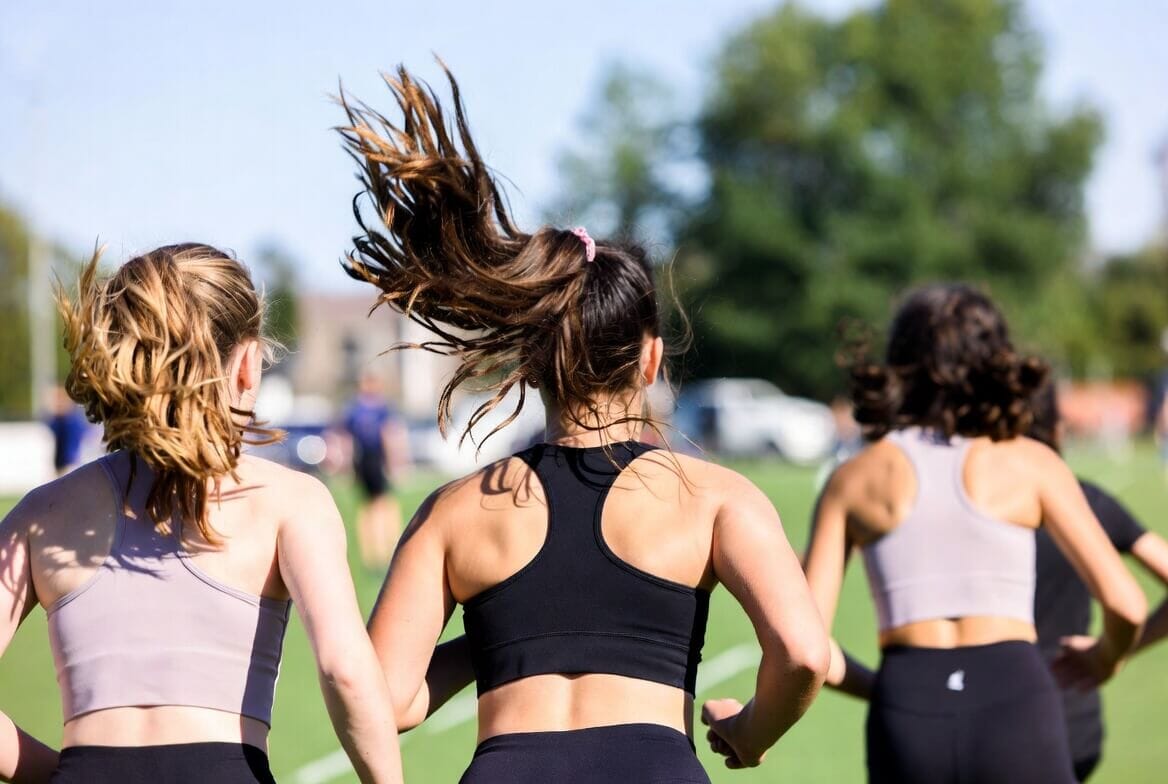 Three young women enjoying outdoor exercise: one girl with curly hair joyfully jumping high with arms outstretched and a big smile, while two others in sportswear stand nearby on a sunny grassy field.
