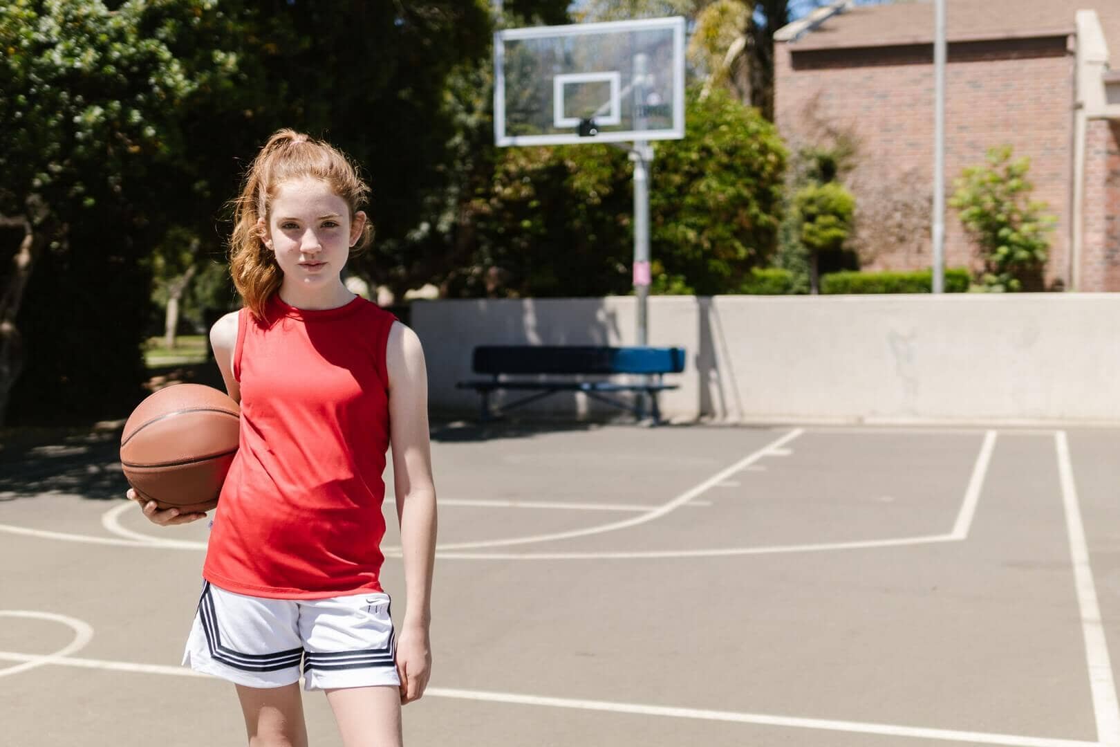 A young girl standing on an outdoor basketball court holding a basketball under her arm. She is wearing a red sleeveless top and white shorts with black stripes.