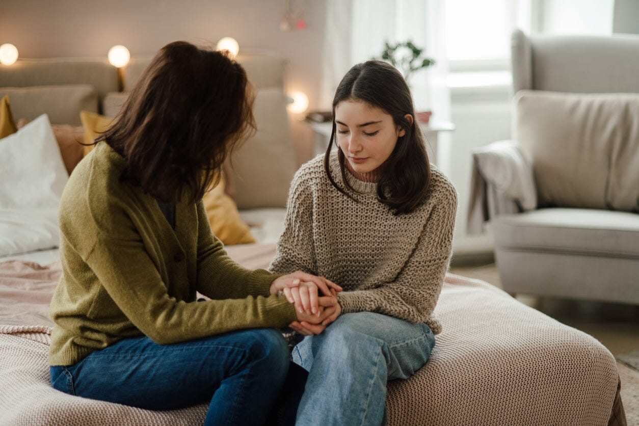 An image of a mother and daughter sitting on a bed holding hands.