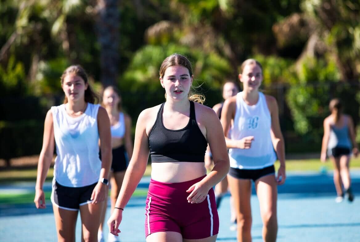 A group of teenage girls in athletic wear walking and running on an outdoor sports court, with one girl in a black sports bra and maroon shorts in the foreground.