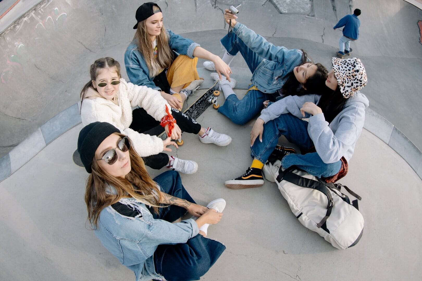 A high-angle fisheye lens shot of a group of six fashionable young women sitting on the ground in a skate park, posing with cameras and backpacks. One girl in the foreground on the left is wearing sunglasses and a black beanie.