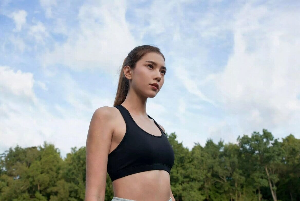 A fit young woman with long brown hair in a ponytail, wearing a black sports bra, standing outdoors in a park with green trees and a blue sky with clouds, looking confidently to the side.