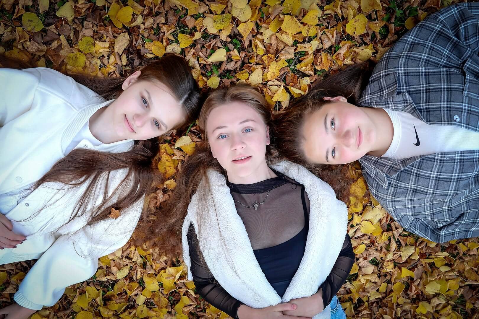 Three young women are seen from a high angle, lying with their heads together on a patch of grass. The grass is heavily covered with fallen, yellow autumn leaves. Each woman has a relaxed expression, looking up towards the camera.