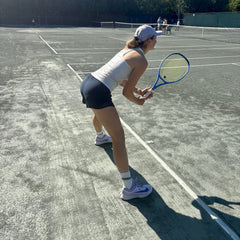 White#A woman on an outdoor tennis court wearing a white tank top, dark shorts, a white cap, and holding a blue and black tennis racket.