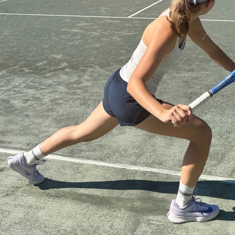 A woman on a tennis court wearing a white tank top, navy athletic skirt, white socks, and light purple Nike sneakers, holding a tennis racket in a dynamic lunge position.