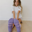 Lilac#A young girl sitting on a light wood floor against a white wall, wearing lavender socks with the text 'THESE FEET DON'T STINK' printed in white on the soles.