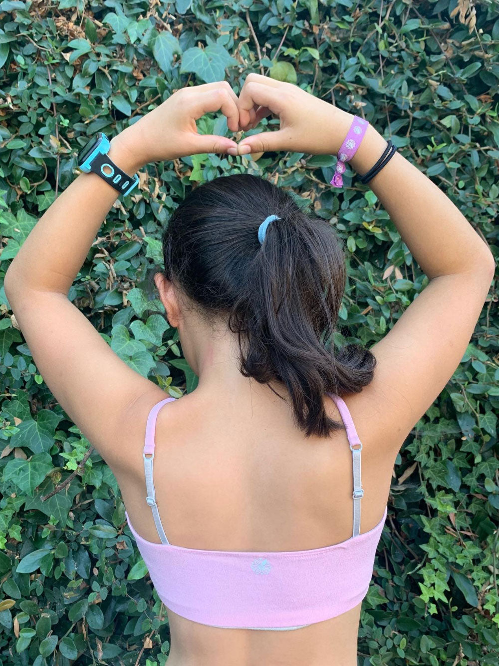 Girl in a ponytail wearing a pink Bleuet Bleum ultra-soft seamless girls first bra and training bra in a garden view from back.  