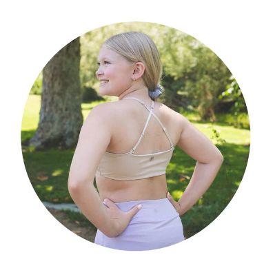 Tween girl in a beige sports bra and purple leggings standing outdoors with trees in the background