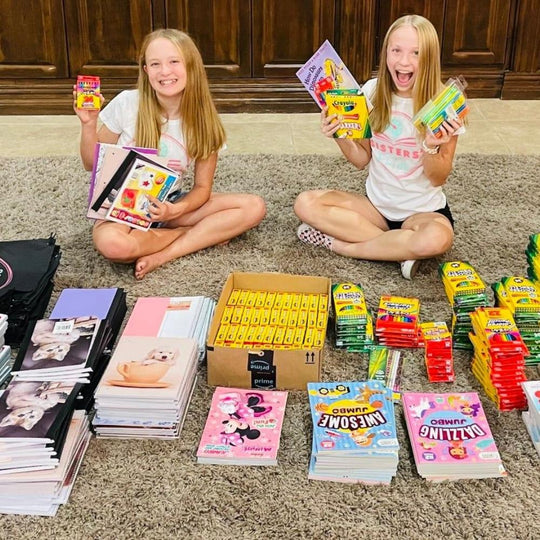 Gift#Two smiling girls sitting on the floor surrounded by various school supplies including notebooks, crayons, markers, and coloring books. They are organizing the items as part of a project named 'Gift of Art for Kids in Need'.