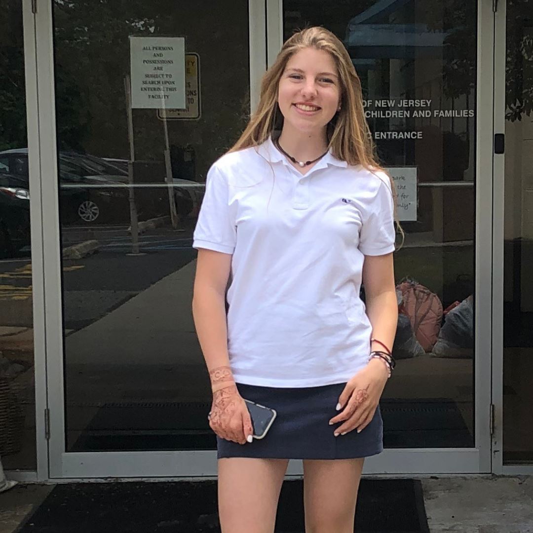 Gift#A girl standing outside in front of a glass door, wearing a white polo shirt and a navy skirt, holding a phone in her right hand.