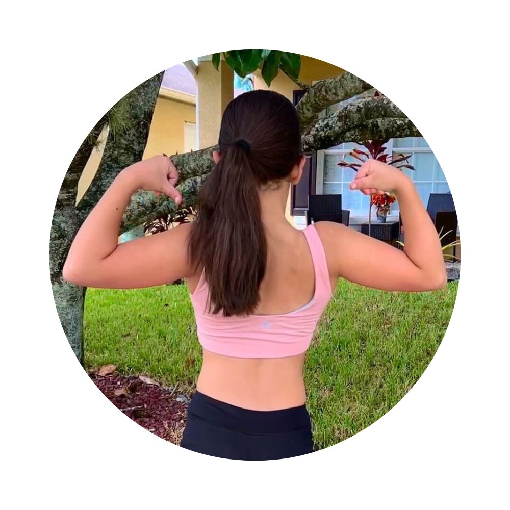 Rose-Mist#A young girl flexing her muscles and wearing a pink Aster Organic Tank Bra outdoors, with greenery and a house in the background.  Rose-Mist#Organic Bras & Bralettes For Girls, Tweens and Teens